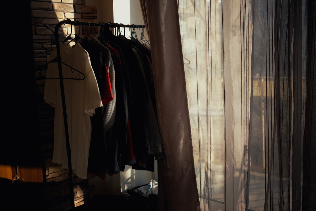 a rack of clothes next to a window with sheer curtains