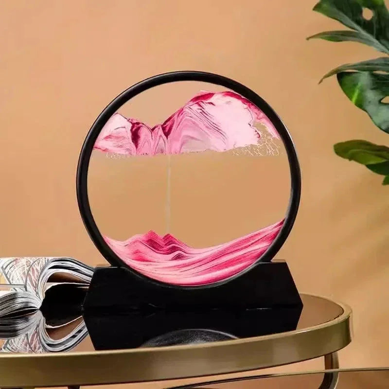Modern round hourglass with pink sand on black stand, placed on reflective gold table with plant and magazine background