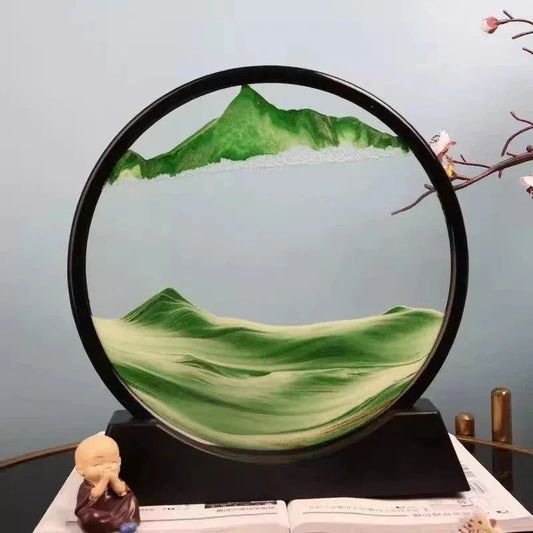 Decorative sand art with green mountains inside a circular black frame on a black base, beside a praying monk figurine