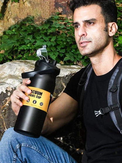 Man outdoors holding black vacuum travel cup with yellow label, sitting on rock, wearing black shirt and backpack